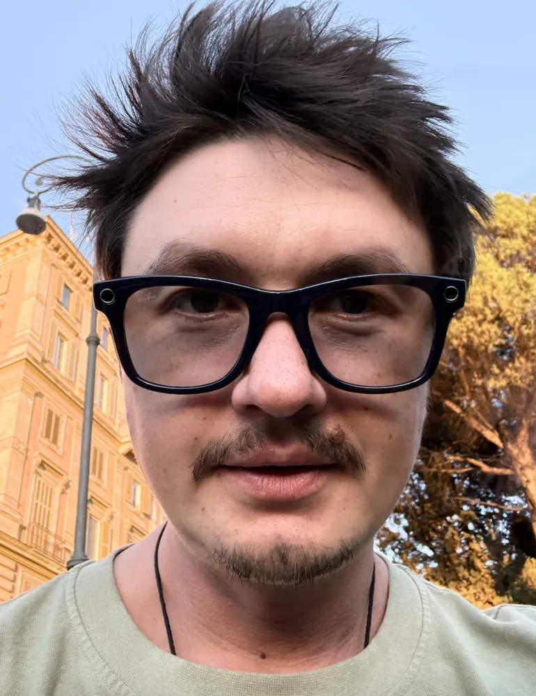 Booga, host of Vibe With Me, outdoors, with tousled dark hair, black glasses, and a light green shirt.